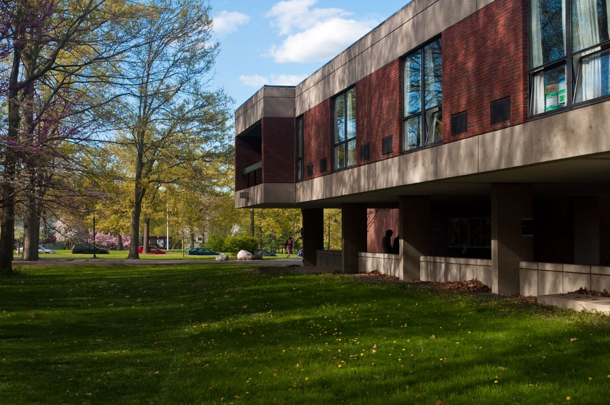 The field next to Franklin Patterson Hall, lush and green, speckled with fallen autumn leaves. Sun and shade dapple the brick brutalist building.